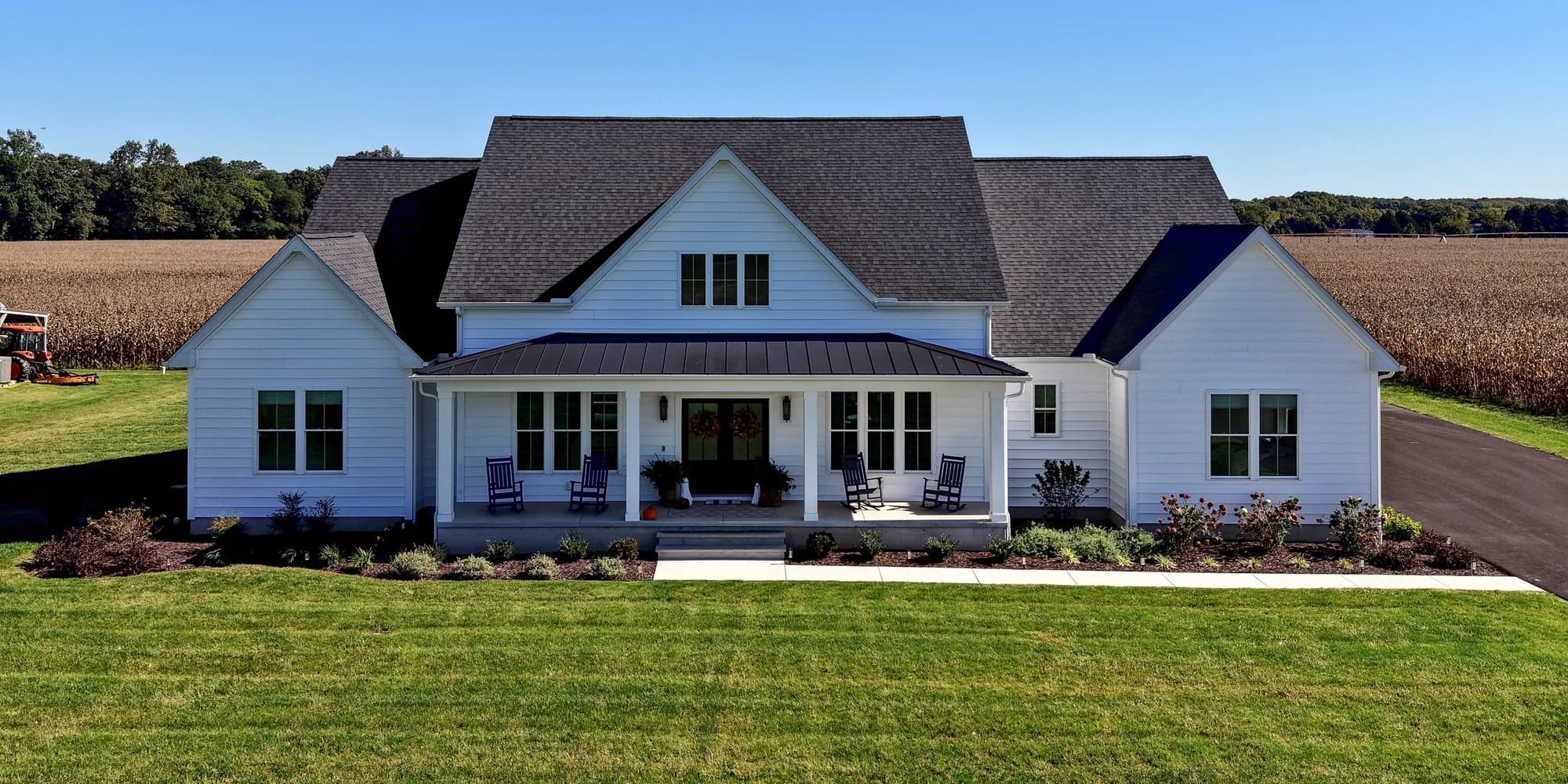 The finished house from the air: a white farmhouse with a dark shingle roof and a metal porch roof, on a wide lawn with corn fields behind it
