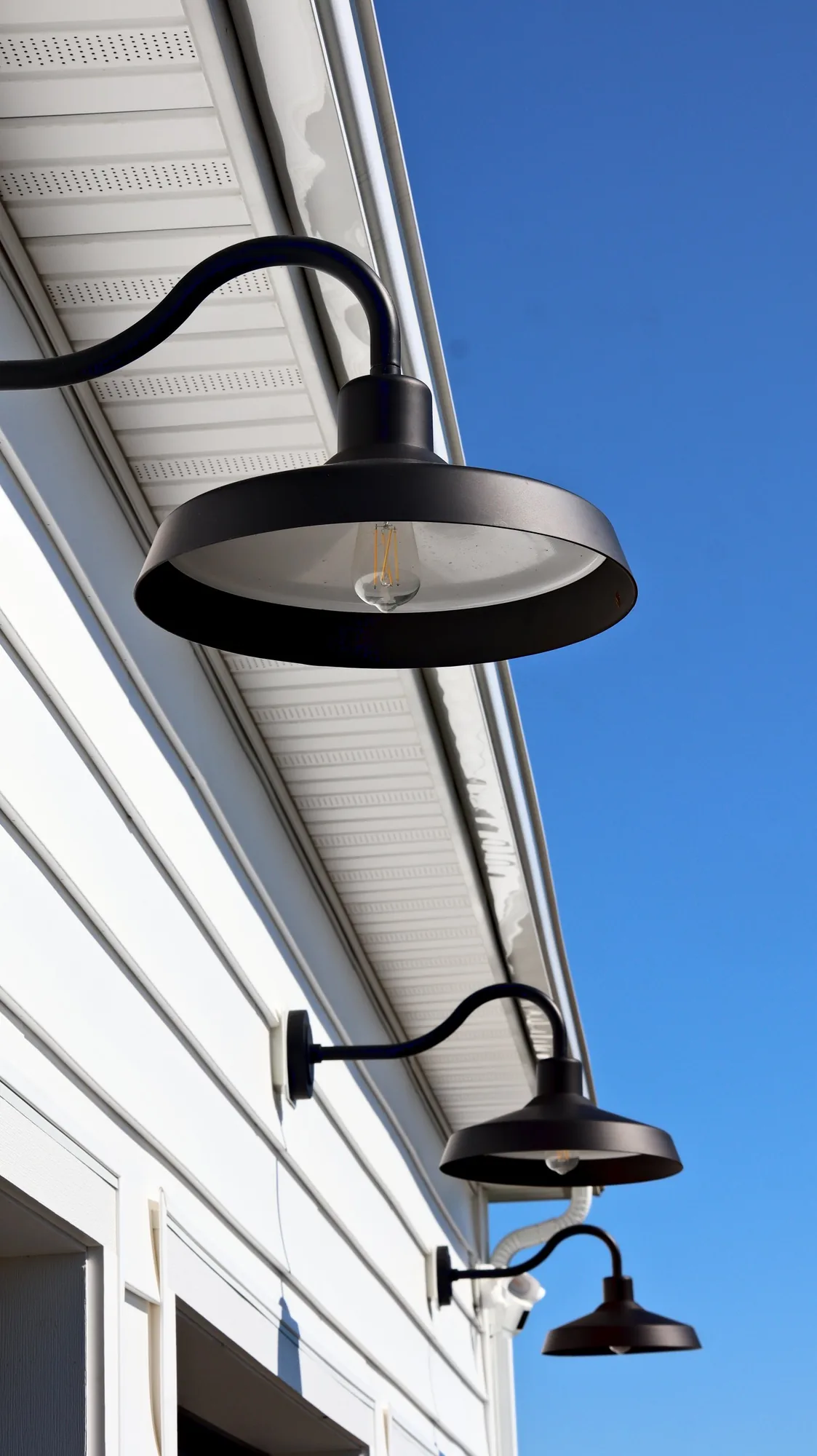 Black gooseneck barn lights on white siding against a clear blue sky
